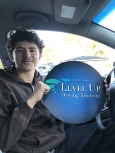 Daniel Garcia, a young man with curly dark hair, sits in a car holding a Level Up Driving Academy sign, smiling confidently after completing his driving lessons.